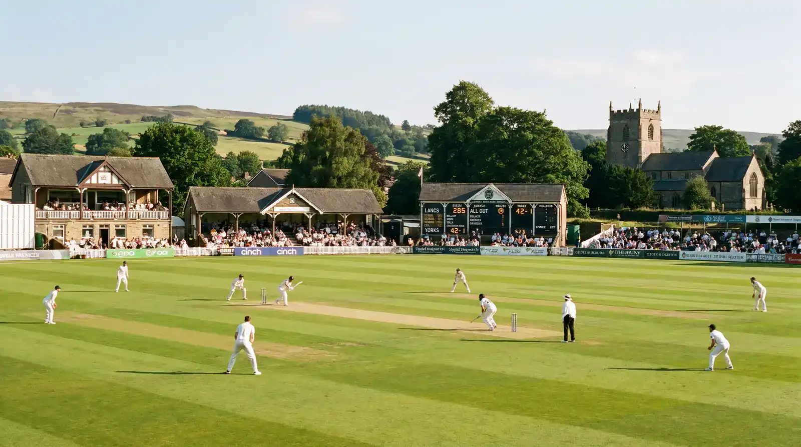 County Championship match in progress at a sunny English ground with a small pavilion crowd and the scoreboard showing first-innings totals