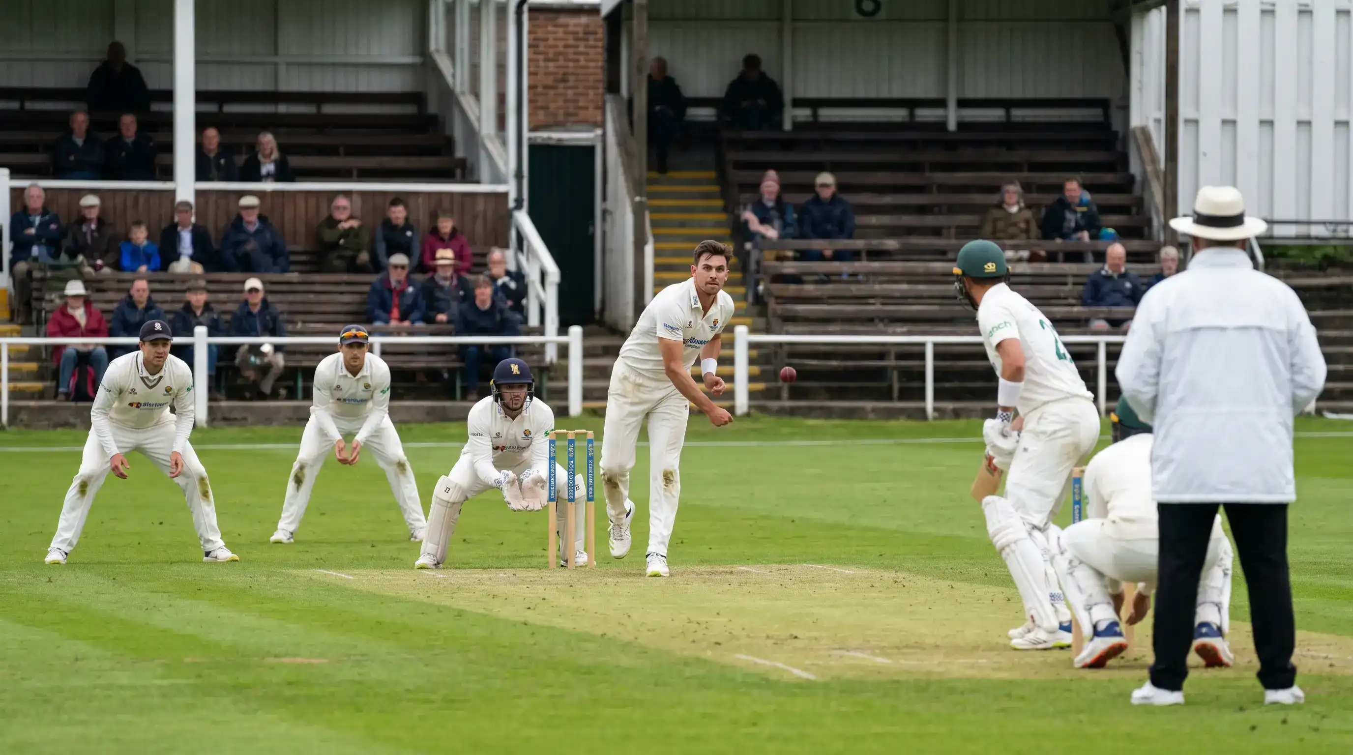 County Championship four-day match in progress with red ball and white kit on a club ground