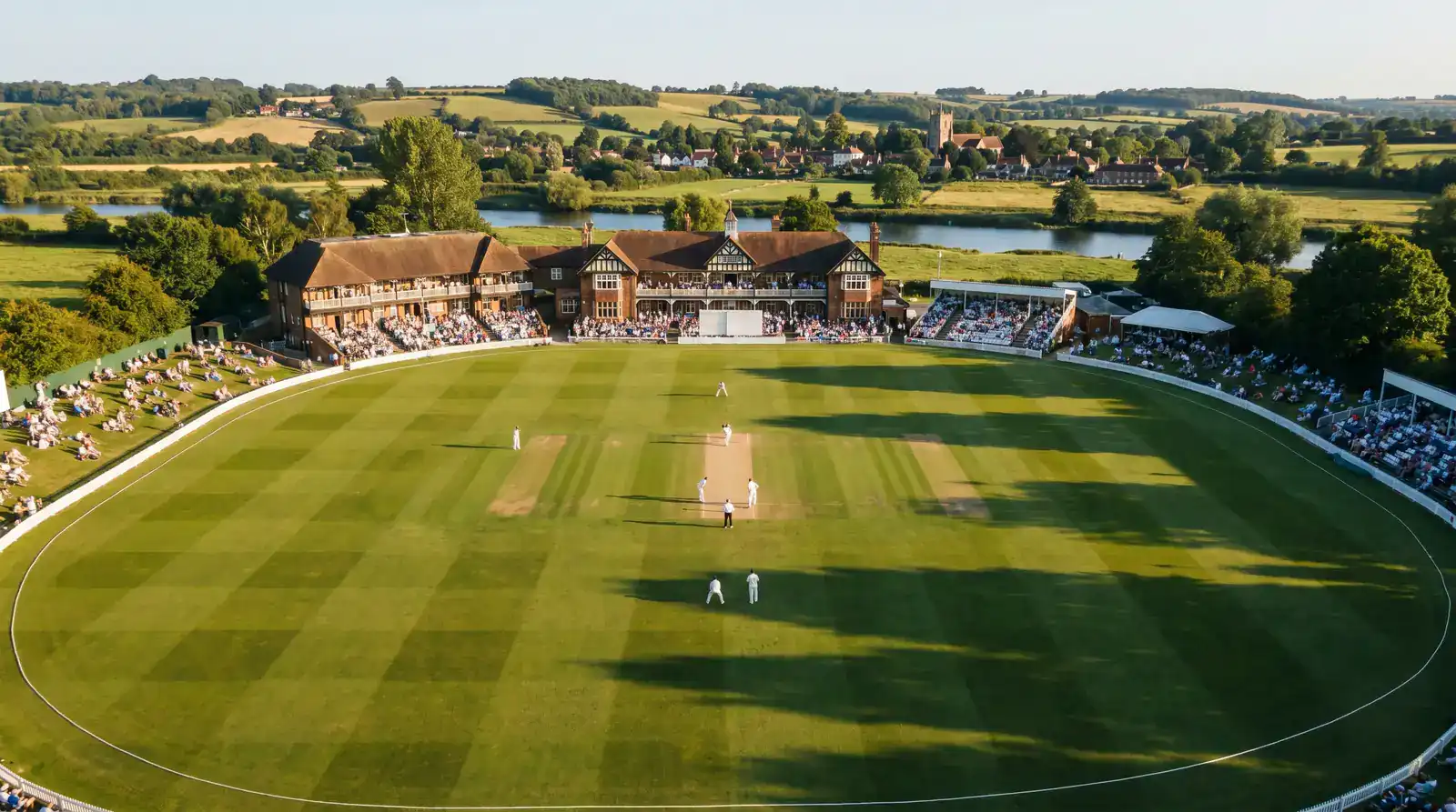 Aerial view of an English cricket ground with pavilion crowd stands and the boundary rope visible on a clear summer afternoon