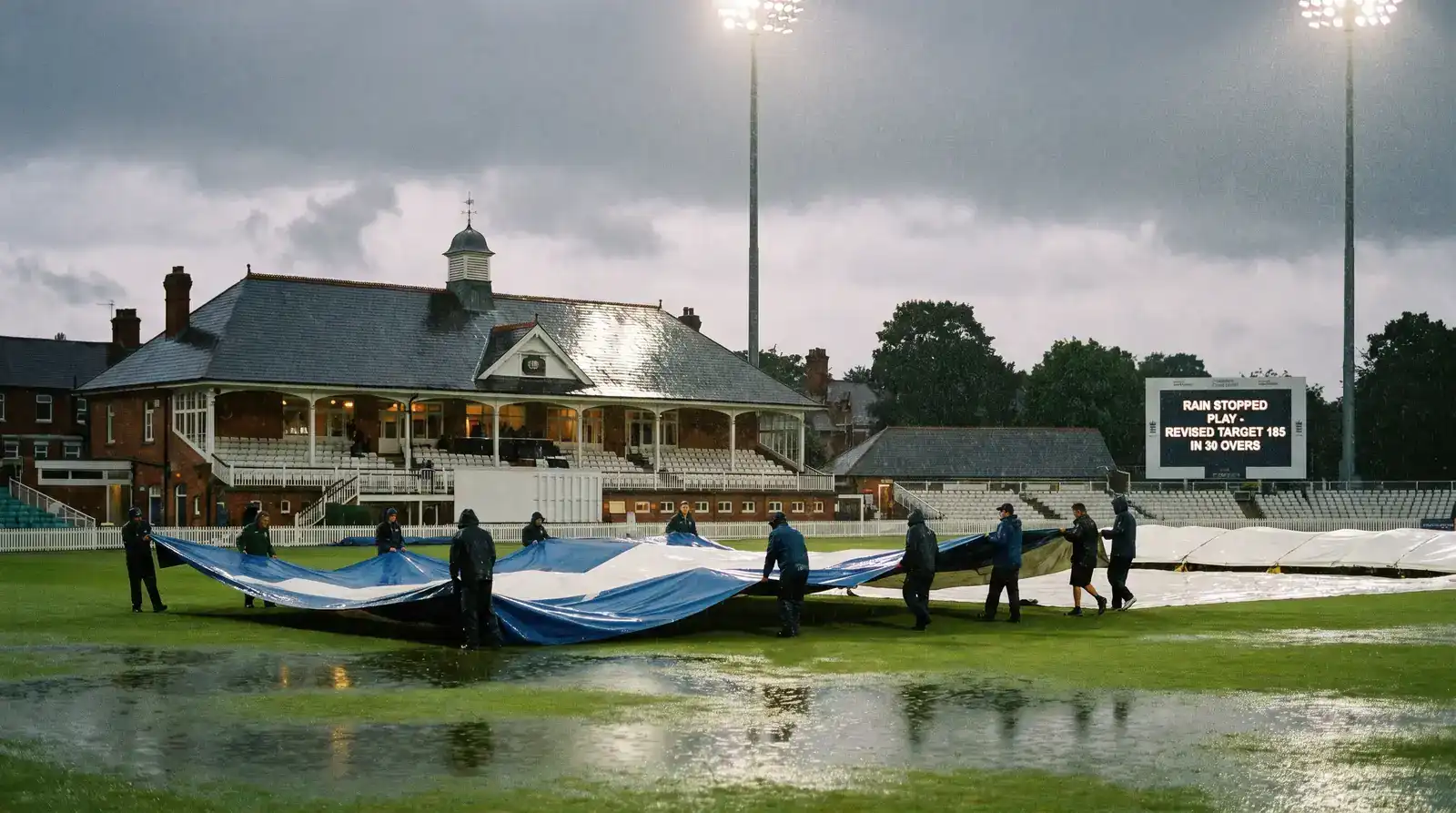 Rain-affected English county cricket ground with the covers being pulled across the square while the electronic scoreboard shows a revised DLS target