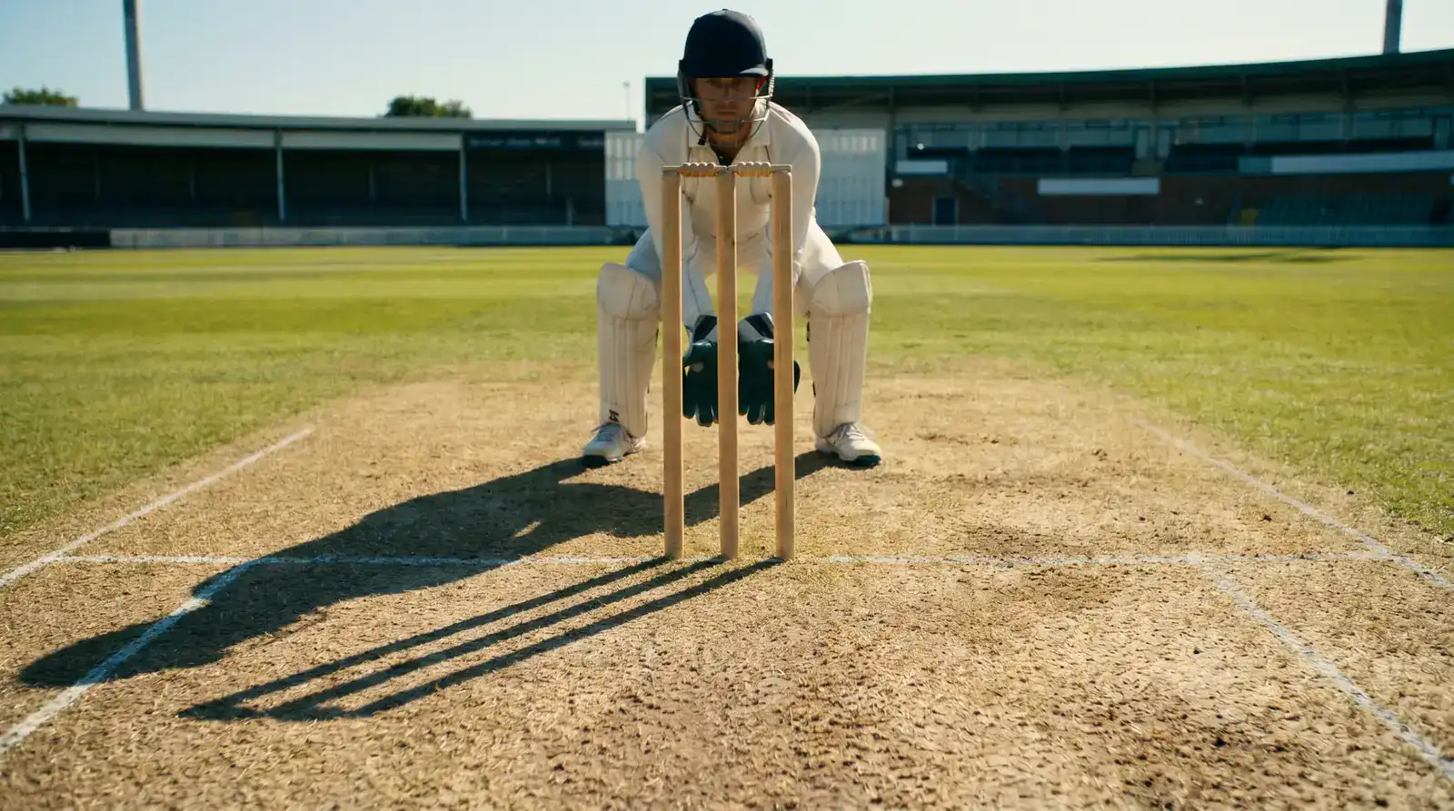 Cricket stumps in clear focus from down the pitch with the wicket-keeper crouched behind, captured at the moment of a delivery