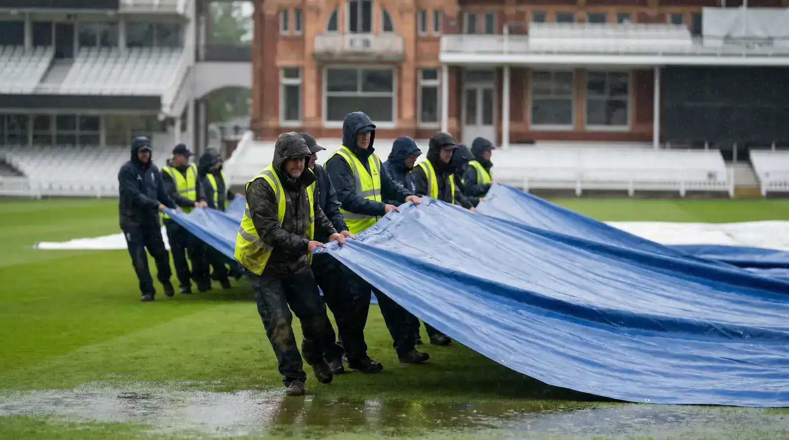 Ground staff pulling rain covers across the pitch during a rain interruption at an English cricket ground