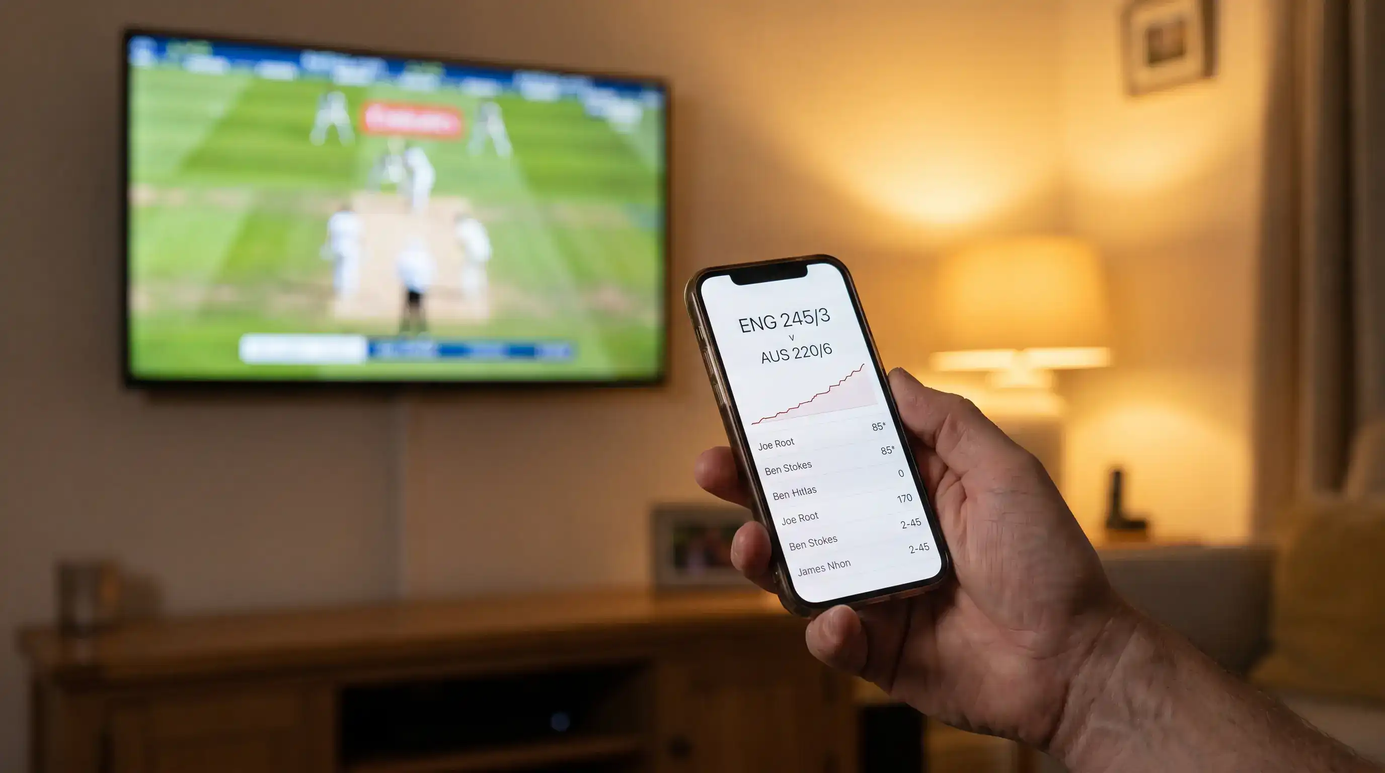 Cricket viewer holding a smartphone showing a live cricket scorecard app while a televised match plays in the background