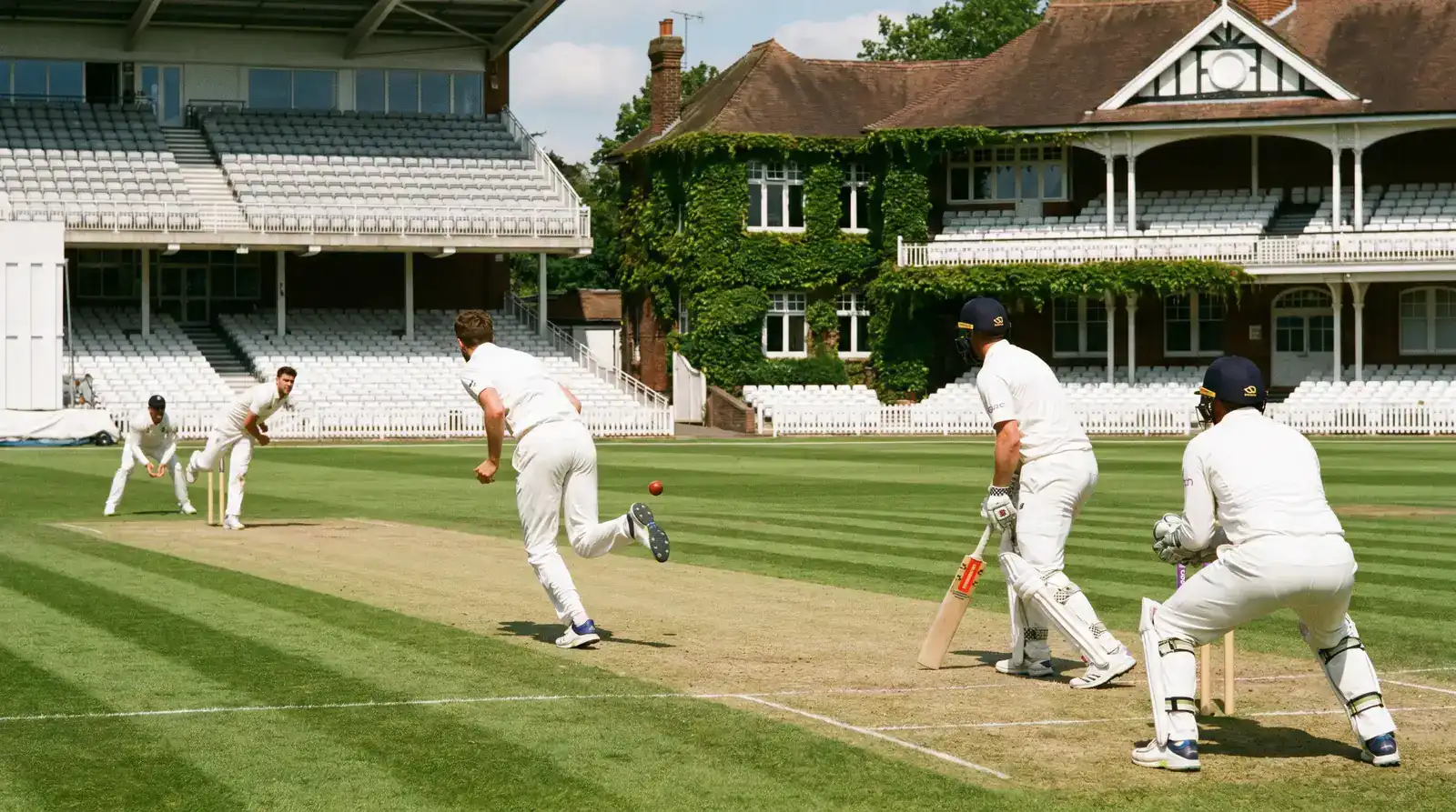 Cricket match in progress on a sunlit English Test-match ground with red-ball whites and a wicket-keeper crouched behind the stumps