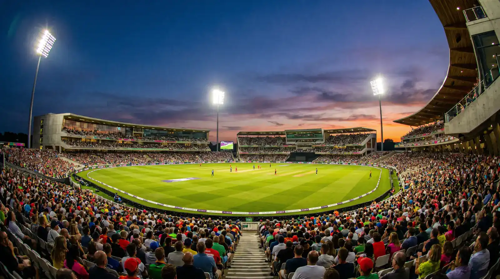 Modern English cricket stadium with floodlights for a Hundred match showing a packed coloured crowd in the upper tiers