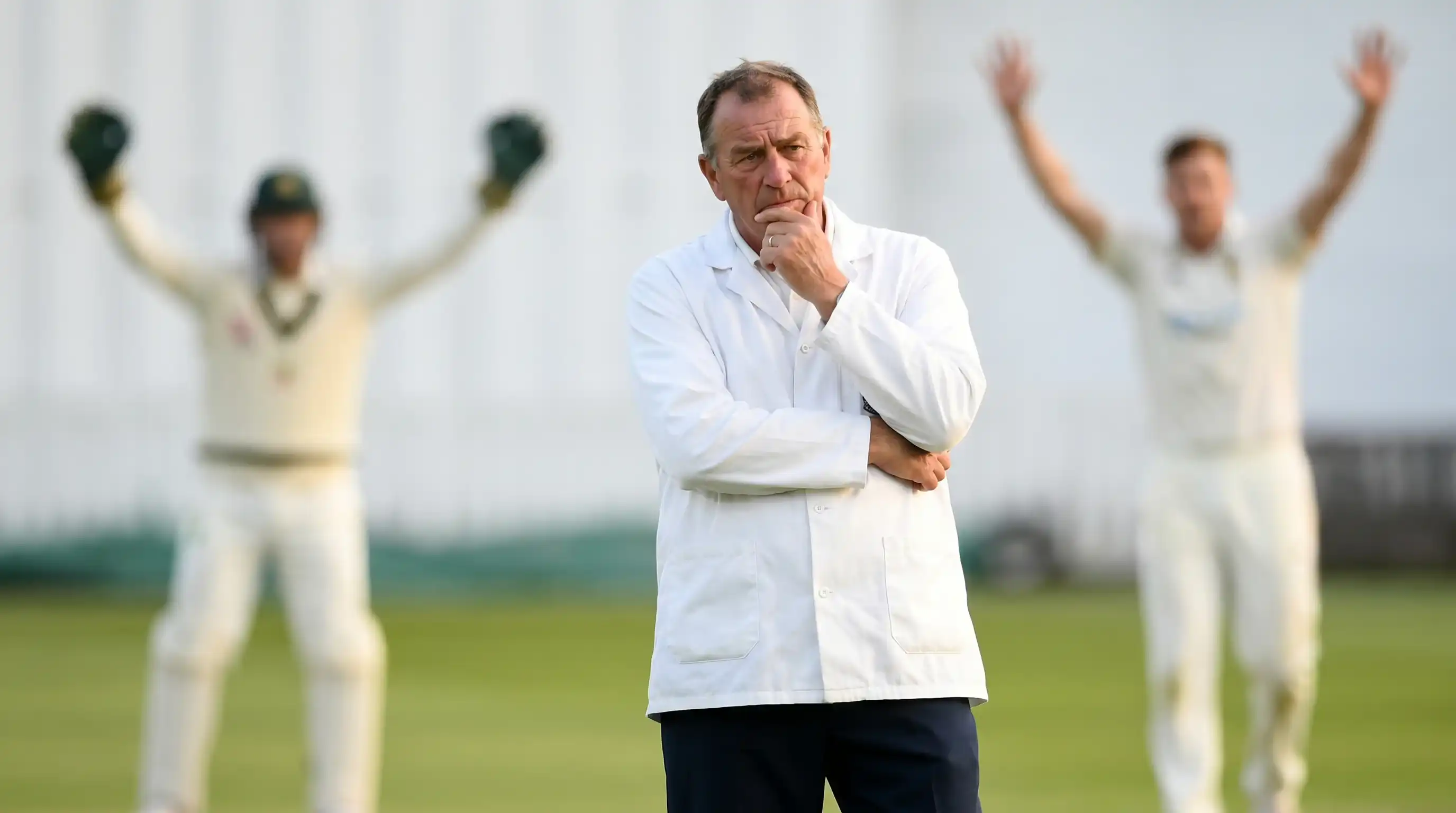 On-field cricket umpire holding his finger thoughtfully on his chin while considering a tight LBW appeal at a county ground