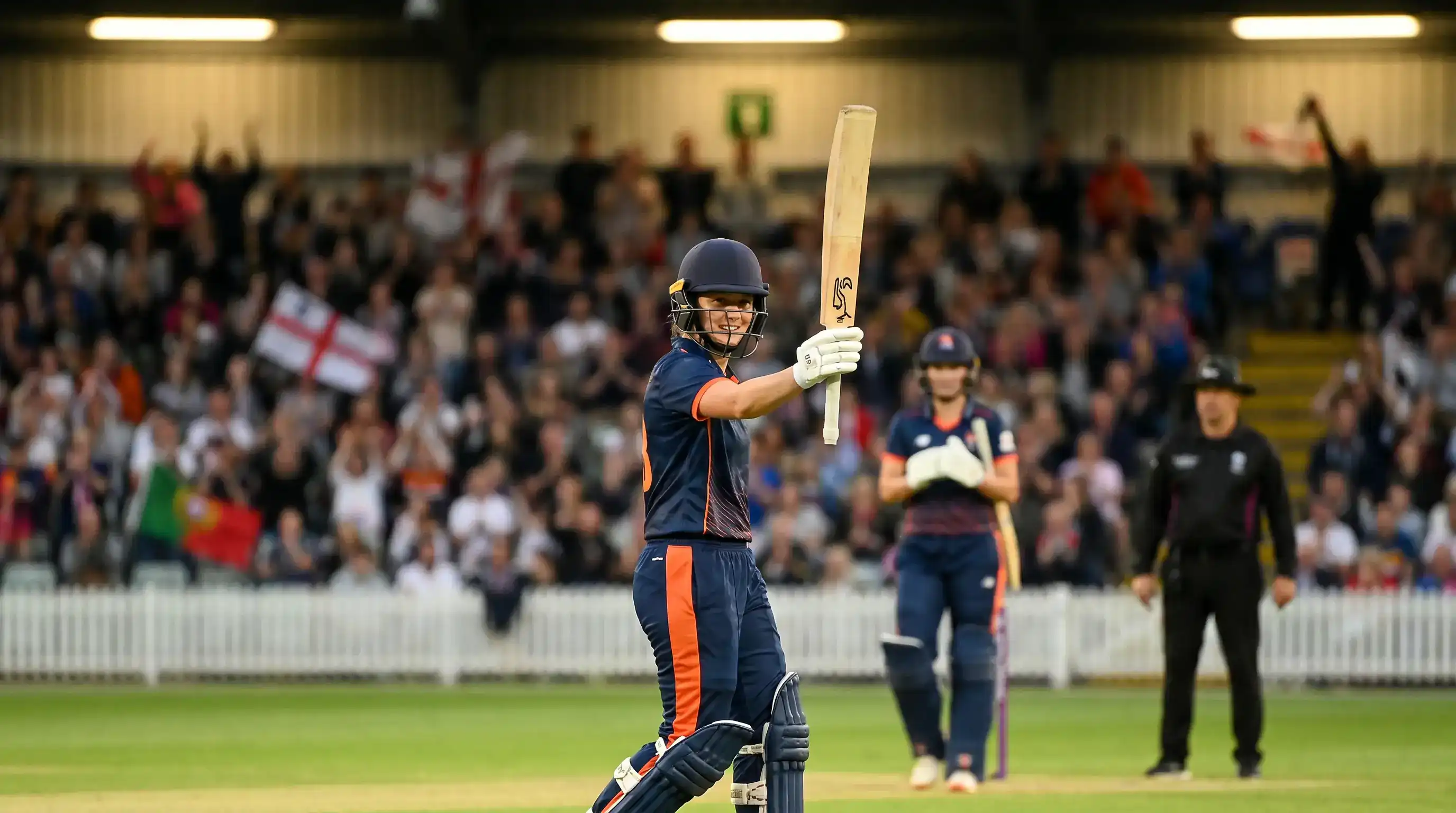 Female cricket batter celebrating a boundary by raising her bat with the crowd applauding in the background