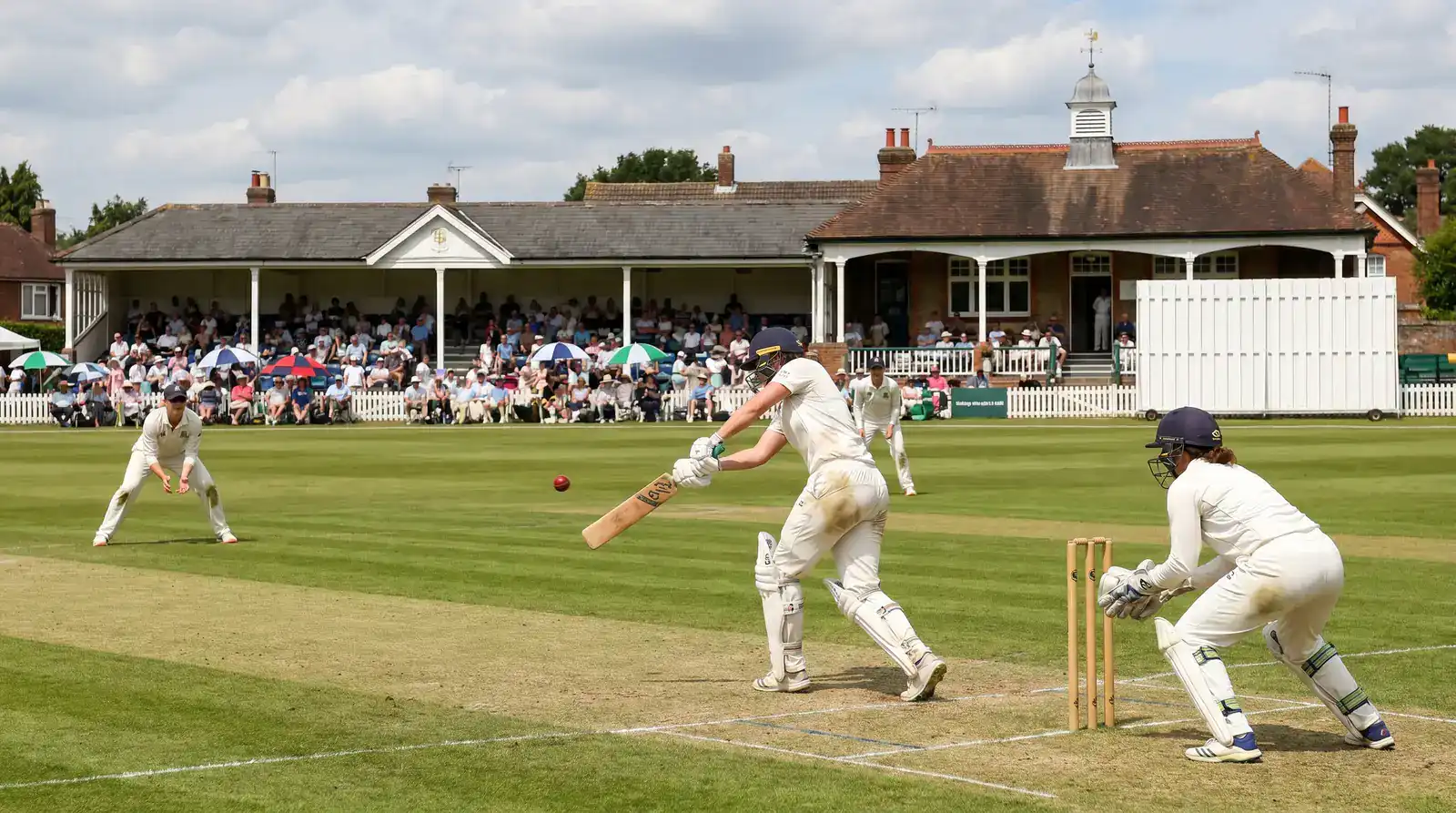 Women's cricket match in progress at a UK ground with a batter playing a drive and the wicket-keeper standing back